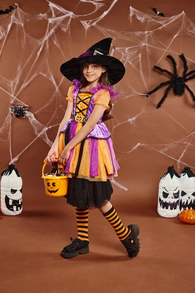 cute preteen in witch hat holds bucket of sweets on backdrop with lanterns and spiders, Halloween