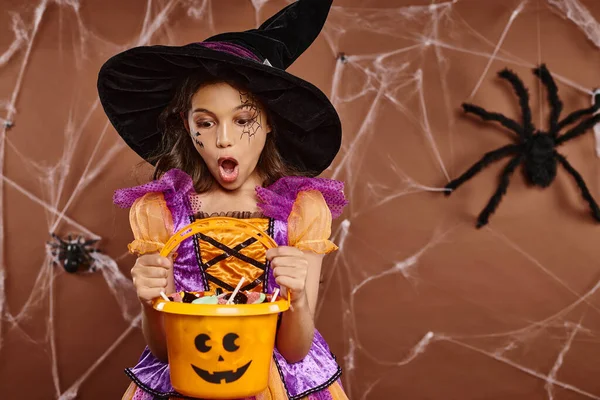 shocked girl in witch hat with spiderweb makeup looking at sweets in Halloween bucket on brown