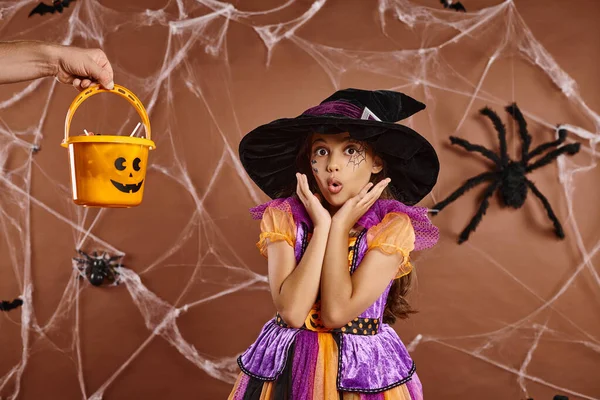 shocked kid in witch hat and Halloween costume looking at camera near hand holding sweets in bucket
