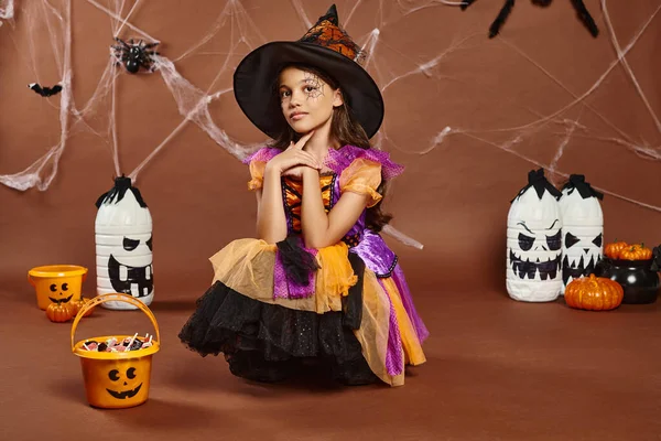 girl in witch hat and Halloween costume looking at camera near sweets in bucket and spooky decor