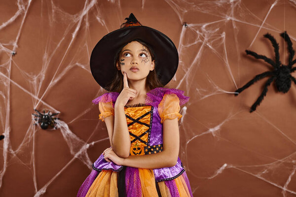 pensive girl in witch hat and Halloween costume standing near cobwebs on brown backdrop, spooky