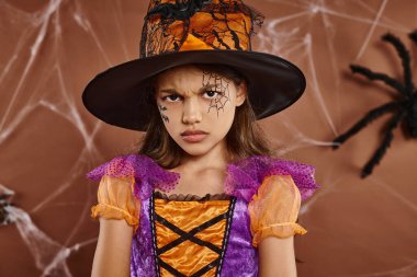 displeased girl in witch hat and Halloween costume frowning on brown background, spooky season clipart