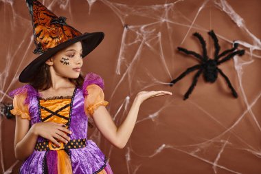 Cadı şapkalı ve Cadılar Bayramı kostümlü kız kahverengi arka planda sahte örümceği işaret ediyor. Ürkütücü sezon.