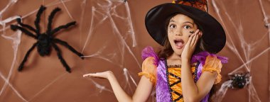 amazed girl in witch hat and Halloween costume pointing at fake spider on brown background, banner clipart