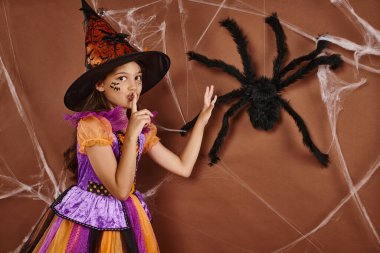 spooky girl in witch hat and Halloween costume showing hush near fake spider on brown background clipart