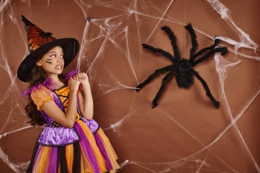 girl in witch hat and Halloween costume grimacing near fake spider and cobwebs on brown background clipart