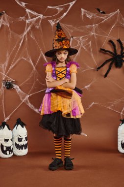 sad girl in witch hat and Halloween costume standing with folded arms on brown backdrop, cobwebs clipart