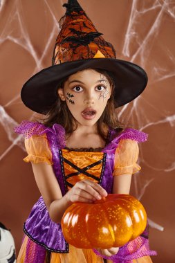 Cadı şapkalı ve cadılar bayramı kostümlü hayret bir kız. Kahverengi arka planda balkabağıyla duruyor. Örümcek ağları.