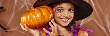 cheerful girl in witch hat and Halloween costume standing with pumpkin on brown backdrop, banner clipart