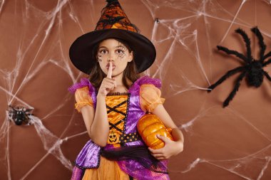girl in witch hat and Halloween costume standing with pumpkin and showing hush on brown background clipart
