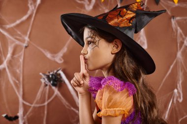 cute little girl in Halloween witch costume and pointed hat showing shh on brown backdrop, side view clipart