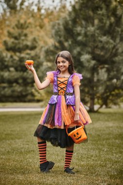 cheerful girl in Halloween costume holding pumpkin and bucket of candies on green grass clipart