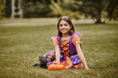 happy girl in Halloween costume sitting in vibrant dress near to decorative pumpkin on green grass