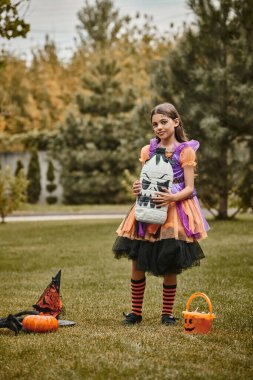 Cadılar Bayramı kostümlü kız balkabağı, sivri şapka ve şeker kovasının yanında tuhaf bir süsleme tutuyor.