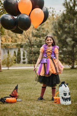 Cadılar Bayramı kostümlü mutlu kız balonları balkabağının yanında, sivri uçlu şapka ve çimlerin üzerinde şeker kovası tutuyor.
