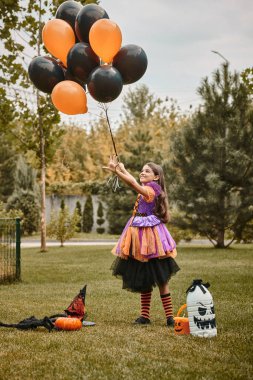 joyful girl in Halloween costume holding balloons near pumpkin, witch hat and candy bucket on grass clipart