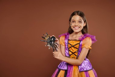 joyous girl in Halloween costume with spiderweb makeup holding fake spider on brown backdrop, spooky clipart