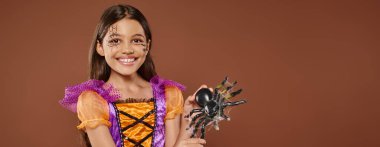 joyous girl in Halloween costume with spiderweb makeup holding fake spider on brown backdrop, banner clipart