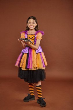 cheerful girl in Halloween costume holding fake spider and standing on brown backdrop, spooky season clipart