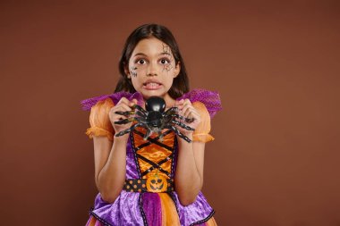 spooky girl in Halloween costume holding fake spider and grimacing on brown backdrop, October 31 clipart