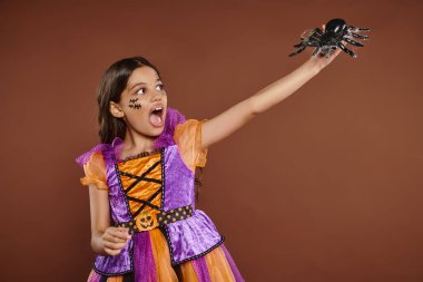 scared girl in Halloween costume looking at black spider and screaming on brown backdrop, October 31 clipart