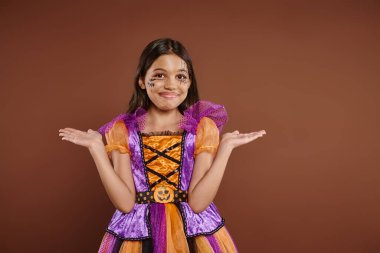 confused girl in Halloween costume with spiderweb makeup smiling and gesturing on brown backdrop clipart