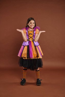 funny girl in Halloween costume with spiderweb makeup smiling and gesturing on brown backdrop clipart