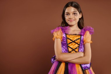 Cadılar Bayramı kostümlü, örümcek ağı makyajlı, kahverengi arka planda katlanmış kolları olan mutlu bir kız.