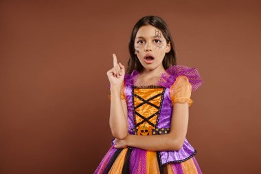 amazed girl in Halloween costume with spiderweb makeup having idea, pointing up on brown backdrop clipart