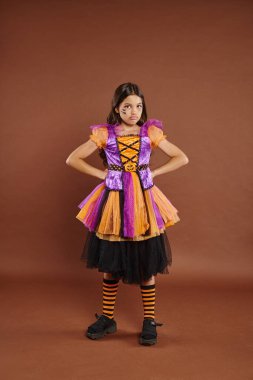 displeased girl in Halloween dress costume standing with hands on hips on brown backdrop, October 31 clipart