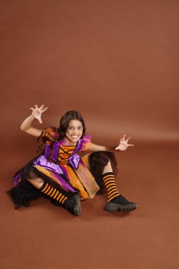 spooky girl in Halloween witch costume sitting and growling on brown backdrop, October 31 clipart