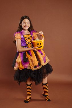 happy girl in colorful Halloween attire holding bucket with candies  and standing on brown backdrop clipart