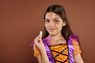 cheerful girl in Halloween costume holding sweet jelly candy on brown backdrop, trick or treat clipart