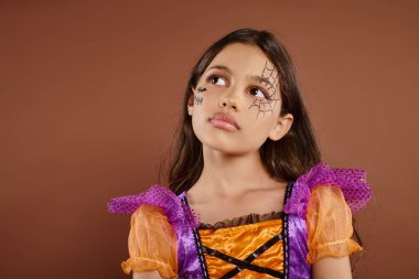 pensive girl in colorful costume with Halloween makeup looking away on brown background, October clipart