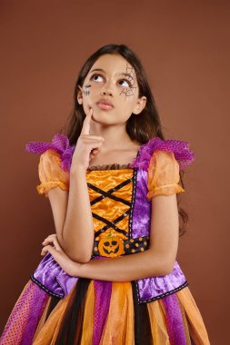 Cadılar Bayramı makyajlı, renkli kostümlü düşünceli bir kız. Kahverengi arka planda, ekim ayında.
