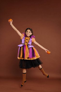 happy girl in Halloween costume levitating with pumpkins on brown background, magic concept clipart