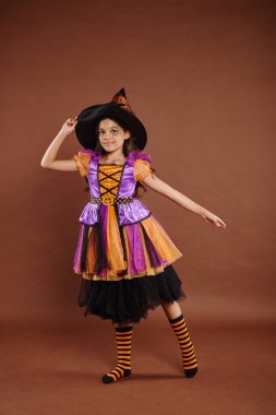 Cadılar Bayramı kostümlü zarif bir kız ve kahverengi arka planda poz veren sivri uçlu bir şapka, mutlu küçük cadı.