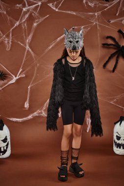 girl in werewolf costume with mask and black faux fur jacket standing on brown backdrop with cobweb