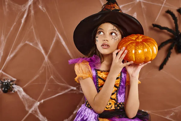 戴着女巫帽穿着万圣节服装站在棕色背景的南瓜上 戴着蜘蛛网的恐怖女孩 — 图库照片