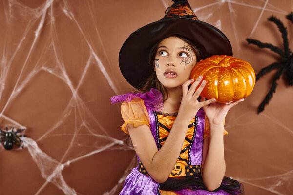 spooky girl in witch hat and Halloween costume standing with pumpkin on brown backdrop, cobwebs