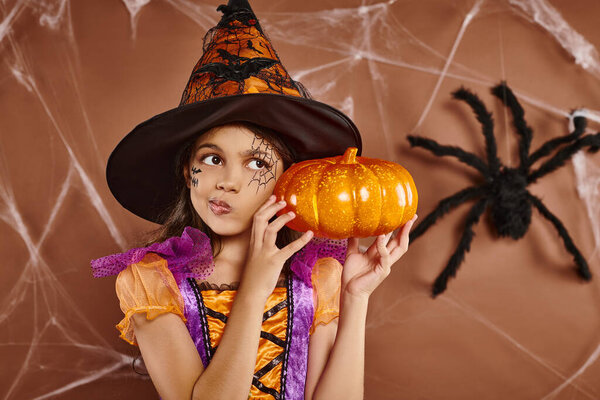 confused girl in witch hat and Halloween costume standing with pumpkin on brown backdrop, cobwebs