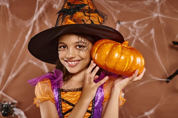 stock image cheerful girl in witch hat and Halloween costume standing with pumpkin on brown backdrop, cobwebs