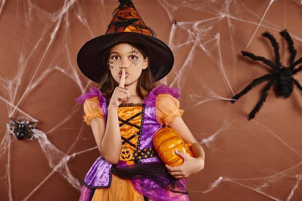 girl in witch hat and Halloween costume standing with pumpkin and showing hush on brown background