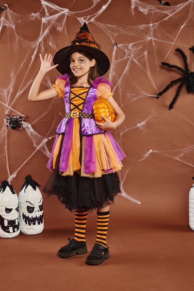 joyful girl in witch hat and Halloween costume standing with pumpkin and waving hand on brown