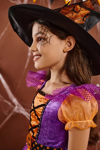 stock image cute little girl in Halloween witch costume and pointed hat smiling on brown backdrop, side view