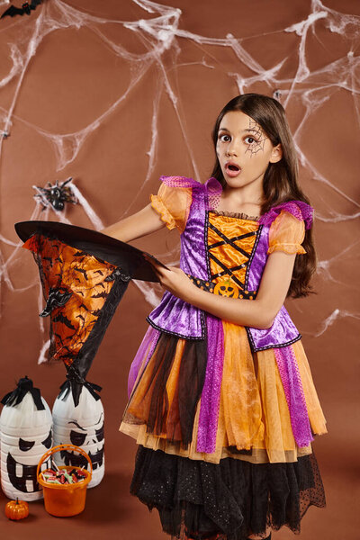shocked child in Halloween witch costume holding pointed hat on brown background, spooky season