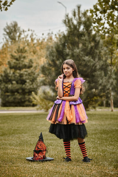 cute girl in Halloween costume standing with lollipop in mouth near pointed hat on green grass