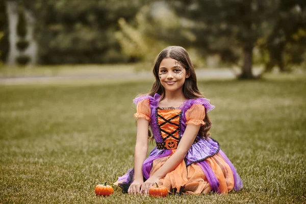 stock image cheerful girl in Halloween costume sitting in vibrant dress near to tiny pumpkins on green grass