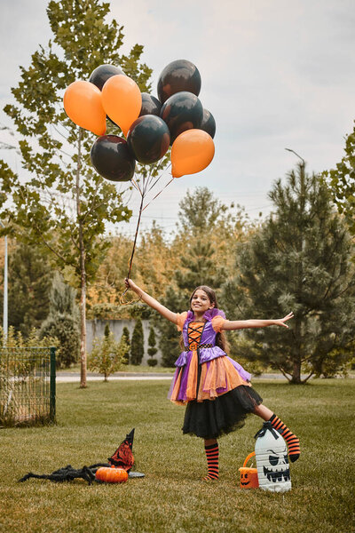 cheerful girl in Halloween dress holding balloons near pumpkin, witch hat and candy bucket on grass