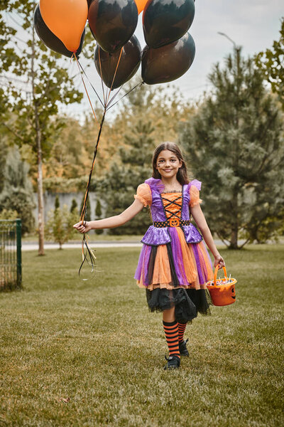 cheerful girl in Halloween costume holding balloons and candy bucket while walking on green grass